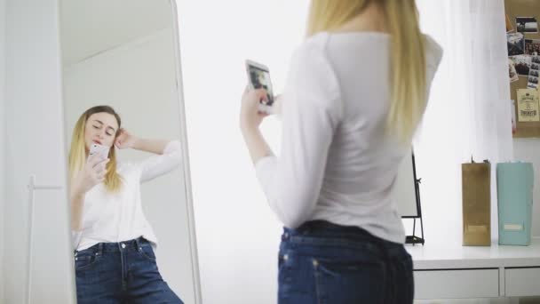 Uma jovem mulher se olha no espelho, se prepara, tira fotos de si mesma em seu telefone no espelho, ajusta suas roupas e penteado. Ele sorri e gosta de si mesmo. — Vídeo de Stock