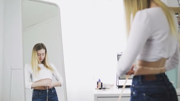 Uma jovem mulher fica na frente de um espelho e mede a circunferência da cintura com uma fita métrica. A menina é complexa por causa do peso — Vídeo de Stock