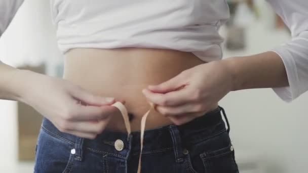 Una mujer joven se para frente a un espejo y mide la circunferencia de su cintura con una cinta métrica. Primer plano — Vídeos de Stock