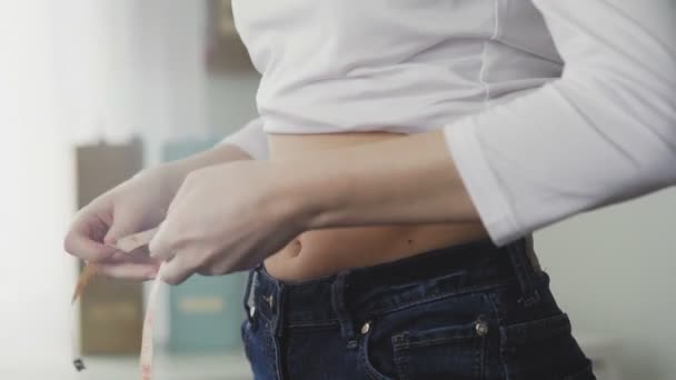 Uma jovem mulher fica na frente de um espelho e mede a circunferência da cintura com uma fita métrica. Close-up — Vídeo de Stock