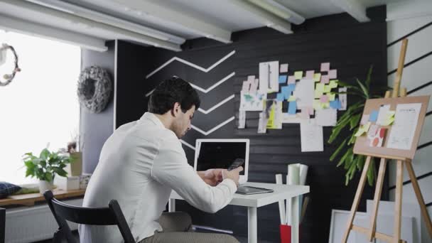 Un joven de apariencia caucásica se sienta en una mesa y trabaja por teléfono. Un joven trabajando remotamente — Vídeo de stock