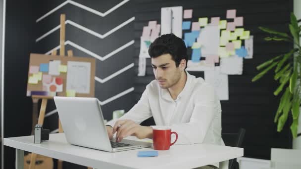 Молодой кавказский человек сидит за компьютером и работает дистанционно, делая заметки на бумаге. Молодой человек, работающий удаленно — стоковое видео