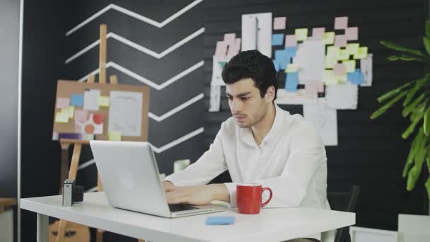 Молодой кавказский человек сидит за компьютером и работает дистанционно, делая заметки на бумаге. Молодой человек, работающий удаленно, пьет кофе или чай. — стоковое видео