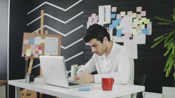 Un joven de apariencia caucásica está sentado en una computadora y trabajando remotamente, tomando notas en papel. Un joven trabajando remotamente — Vídeo de stock