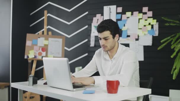 Un joven de apariencia caucásica está sentado en una computadora y trabajando remotamente. El joven coge el teléfono y escribe un mensaje — Vídeo de stock