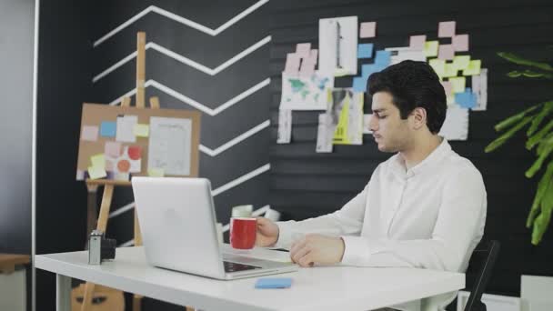 Un joven de apariencia caucásica está sentado frente a una computadora mirando por la ventana y bebiendo té o café. El joven trabaja remotamente. — Vídeo de stock