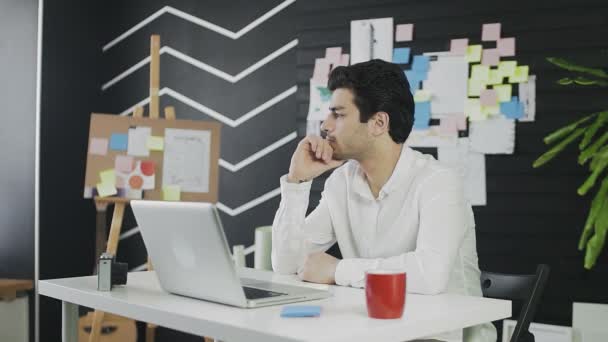 Un joven reflexivo de apariencia caucásica está sentado en una computadora y trabajando remotamente. El joven trabaja remotamente. — Vídeos de Stock