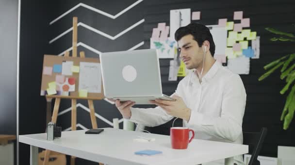 Un joven de apariencia caucásica está sentado en una computadora y trabajando remotamente. Un joven con auriculares se comunica a través de un enlace de vídeo y muestra sus notas en la pared — Vídeo de stock