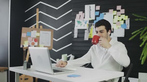 Un joven de apariencia caucásica está sentado en una computadora y trabajando remotamente. Un joven con auriculares se comunica a través de un enlace de vídeo y bebe té o café — Vídeo de stock