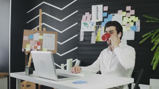 Молодой кавказский человек сидит за компьютером и работает удаленно. Молодой человек в наушниках заканчивает видеоразговор и пьет чай или кофе — стоковое видео