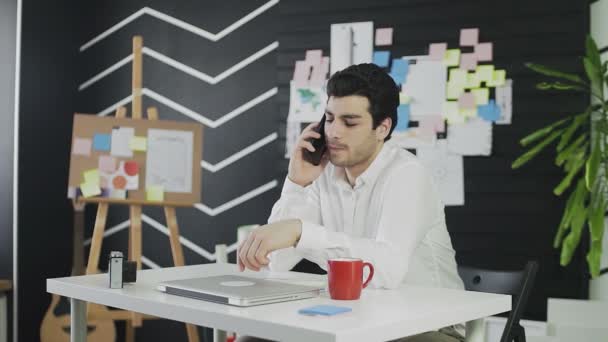 Un joven de apariencia caucásica está sentado en una mesa y hablando por teléfono. El joven trabaja remotamente. — Vídeo de stock