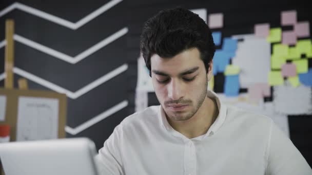 Крупный план молодого человека кавказской внешности, сидящего за компьютером и работающего дистанционно, делающего заметки на бумаге. Молодой человек, работающий удаленно. Видео в движении — стоковое видео