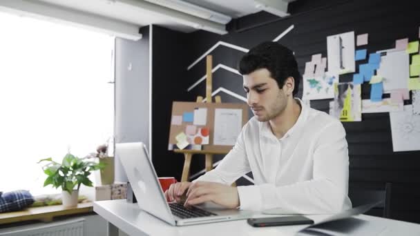 Een blanke jongeman zit achter een computer. Op afstand werken. Freelance. Video in beweging — Stockvideo
