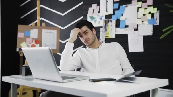 Задумчивый молодой человек с кавказской внешностью сидит за компьютером. Молодой человек работает дистанционно. Видео в движении. Фрилансер. Приближается к камере — стоковое видео