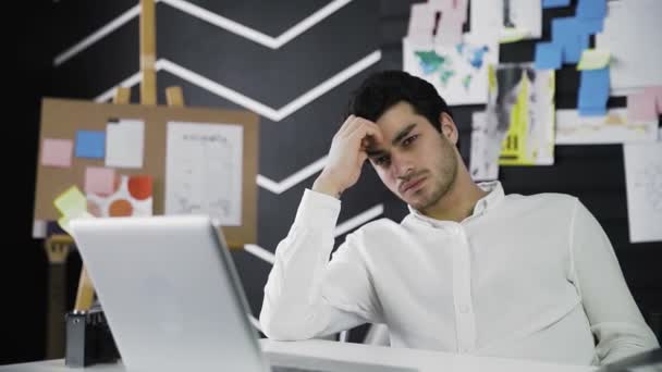 Un joven reflexivo de apariencia caucásica está sentado en una computadora. El joven trabaja remotamente. Video en movimiento. Independiente. Acercamiento de la cámara — Vídeo de stock