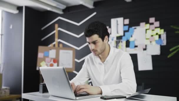 Een blanke jongeman zit achter een computer naar de camera te kijken. Op afstand werken. Freelance. Inzoomen op de camera — Stockvideo