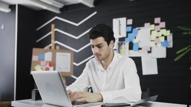 En ung mand af kaukasisk udseende sidder ved en computer og ser vredt på kameraet. Fjernarbejde. Freelance. Zoomer ind på kameraet – Stock-video