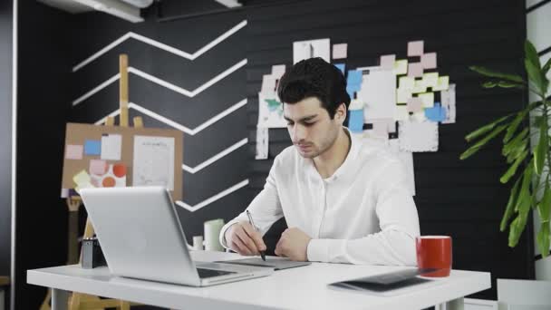 Un joven de apariencia caucásica se basa en una tablilla gráfica. Trabajo remoto. Independiente. Video en movimiento — Vídeo de stock