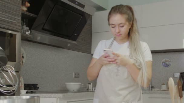 Uma menina na cozinha está procurando receitas na Internet em seu telefone — Vídeo de Stock