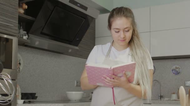 Uma menina na cozinha está à procura de receitas em um livro de receitas ou em um caderno — Vídeo de Stock