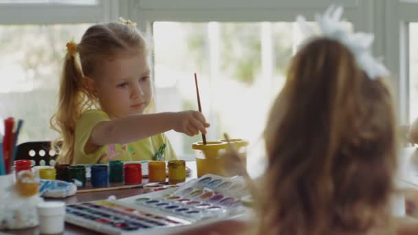 Τα παιδιά κάθονται στο τραπέζι και ζωγραφίζουν σε χαρτί με διαφορετικά χρώματα και πινέλα — Αρχείο Βίντεο