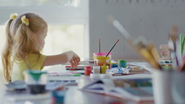 Küçük bir kız çizim için farklı boyalarla dolu bir masada oturuyor. Çocuk çiziyor. Küçük metal bir bardaktaki boya fırçasının yakın çekimi. — Stok video