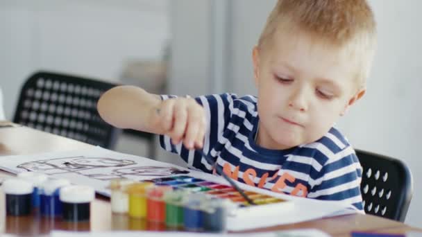 Un bambino è seduto a un tavolo e disegno su carta con colori e pennelli diversi — Video Stock