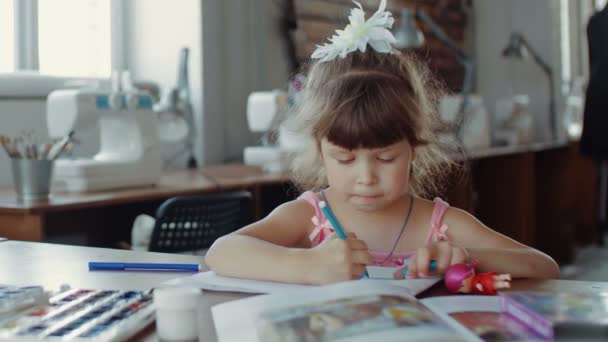 Küçük bir kız masada oturuyor ve farklı renkler ve fırçalarla kağıda resim çiziyor. — Stok video