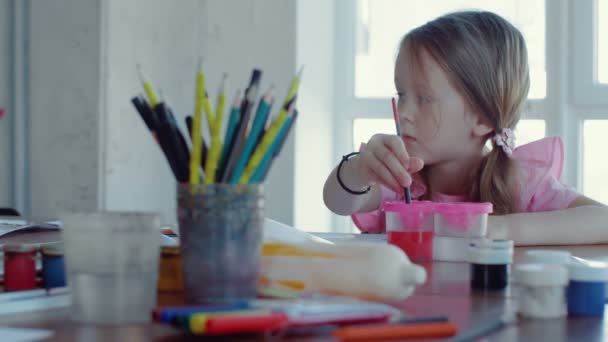 A little girl is sitting at a table and drawing on paper with different colors and brushes — Stock Video