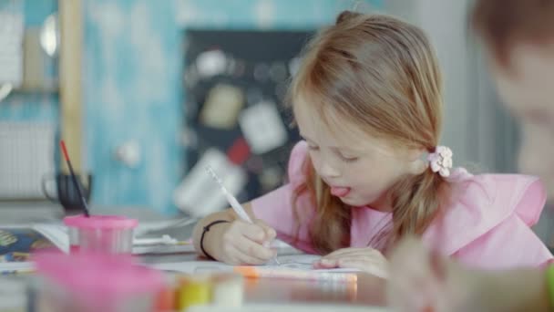 Vista laterale una bambina è seduta a un tavolo e disegna su carta con colori e pennelli diversi — Video Stock