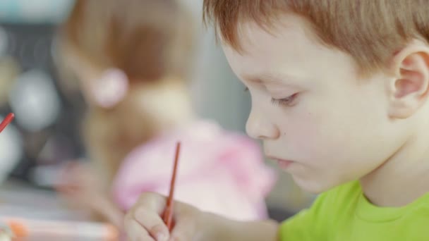 Vista lateral um menino e uma menina estão sentados a uma mesa e desenhando em papel com cores e pincéis diferentes — Vídeo de Stock