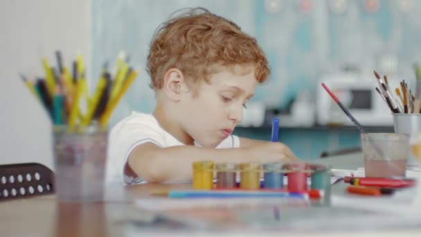 Ein kleiner Junge sitzt an einem Tisch und zeichnet auf Papier mit verschiedenen Farben und Pinseln — Stockvideo