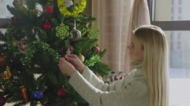 Süveter giyen genç ve güzel bir sarışın Noel ağacının yanında oturuyor ve bir Noel balosu düzenliyor. Evin penceresinin yanında altında hediyeler olan bir Noel ağacı..