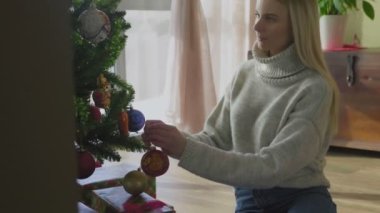 Süveter giyen genç ve güzel bir sarışın Noel ağacının yanında oturuyor ve bir Noel balosu düzenliyor. Evin penceresinin yanında altında hediyeler olan bir Noel ağacı..