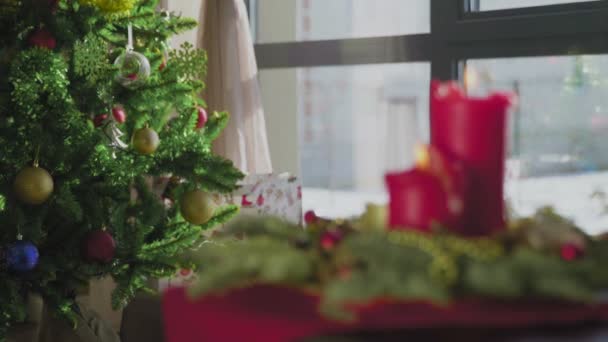 Una corona sempreverde con due candele rosse e una candela dorata. Sullo sfondo, un albero di Natale decorato. Tradizioni e costumi natalizi dei cristiani alla vigilia della festa. — Video Stock