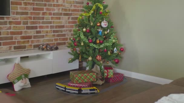 En la esquina de la casa hay un árbol de Navidad con un montón de regalos debajo de él, y un tren de juguete pasea alrededor del árbol. Ambiente acogedor — Vídeos de Stock