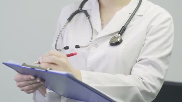 Крупным планом врача женщина в медицинском халате со стетоскопом вокруг шеи, держа в руках таблетку и делая заметки — стоковое видео