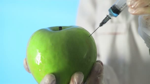 Κοντινό πλάνο ενός ανθρώπου σε ένα ιατρικό παλτό και γάντια που εγχέουν μια σύριγγα σε ένα μήλο με κάποιο υγρό σε μπλε φόντο — Αρχείο Βίντεο