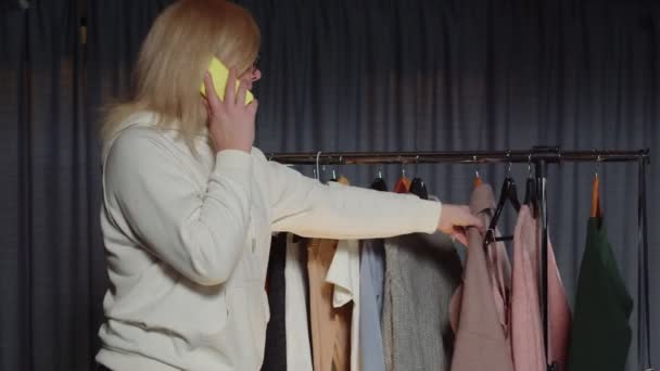 Primer plano de una mujer hablando por un teléfono móvil. Una mujer atractiva está hablando en un teléfono móvil, eligiendo ropa — Vídeos de Stock
