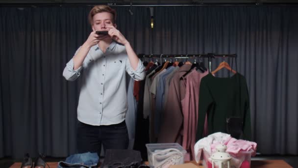 Een jonge vrouw kiest kleren in een winkel en neemt foto 's van hen op haar telefoon. — Stockvideo