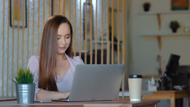 En ung kvinna sitter vid en dator på kontoret, tar ett kaffeglas och kastar det, rätar ut håret och tittar på kameran — Stockvideo