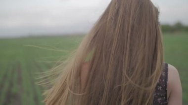 Çiçek baskı ile koyu elbiseli güzel sağlıklı kadın durumda ve yeşil tarlada çalışan geriye bakıp saçını kapatmak kadar yetersiz düz renk esen rüzgar