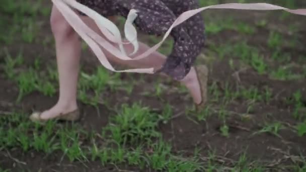 Szép lány szalag sötét ruhában, floral nyomtatási halad át a zöld mező, és a szél fúj neki haj kamera követi a cipők, szoros lövés szembe — Stock videók