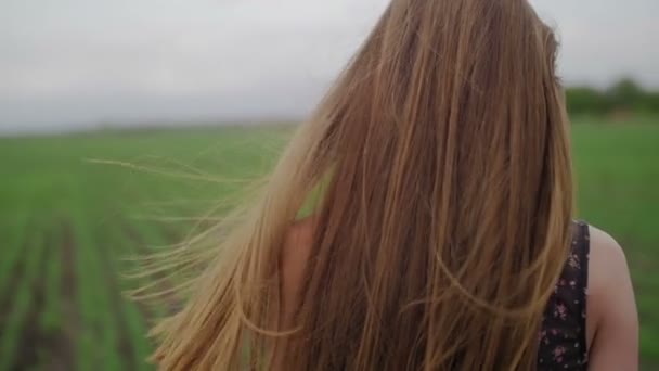 Mulher saudável bonita em vestido escuro com estampas florais fica em e, em seguida, correr através de campo verde olhar para trás e vento soprando seu cabelo de perto — Vídeo de Stock