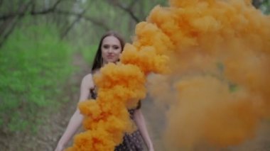 Yaz ormanında açık havada eğlenen çiçek baskılı koyu renkli genç kadın. Renkli turuncu bir sis bombası tutan güzellik kızı ve kameraya gider.harika bir gülümseme. orta çekim yavaş hareket