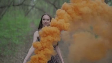 Yaz ormanında açık havada eğlenen çiçek baskılı koyu renkli genç kadın. Renkli turuncu bir sis bombası tutan güzellik kızı ve kameraya gider.harika bir gülümseme. orta çekim yavaş hareket