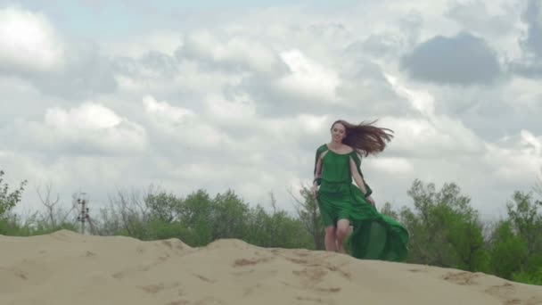 Beauty Girl in green dress running on sand dune while wind blowing her clothes slow motion sky background medium long shot front view — Stock Video