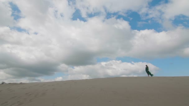 Belleza Chica en vestido verde de pie en la duna de arena, mientras que el viento soplando su ropa cámara lenta cielo fondo largo tiro — Vídeos de Stock