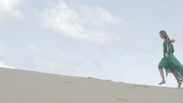 Девушка-красавица в зеленом платье работает на песчаной дюне, в то время как ветер дует ее одежду замедленное движение небо фоне среднего длинный выстрел ungraded плоский цвет — стоковое видео
