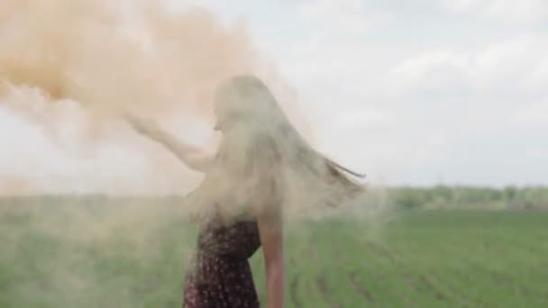 Jovem feliz em vestido escuro com estampa floral se divertindo ao ar livre no campo de geen. Menina beleza com laranja bomba de fumaça girando e rindo. tiro do meio — Vídeo de Stock